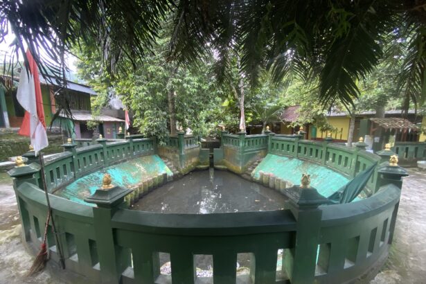 Circular green fountain with a decorative railing and gold finials set in a tropical courtyard, trees overhead and an Indonesian flag nearby