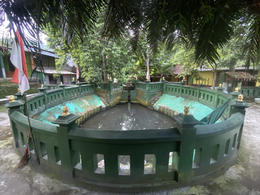 IMG_7295 - Mabur.co Circular green fountain with a decorative railing and gold finials set in a tropical courtyard, trees overhead and an Indonesian flag nearby