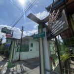 Sunlit urban street with overhead power lines, a green fence, and shop signs in Indonesian.