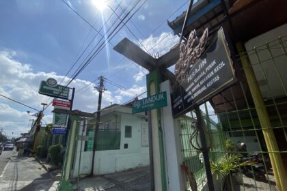 Sunlit urban street with overhead power lines, a green fence, and shop signs in Indonesian.