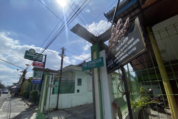 Sunlit urban street with overhead power lines, a green fence, and shop signs in Indonesian.