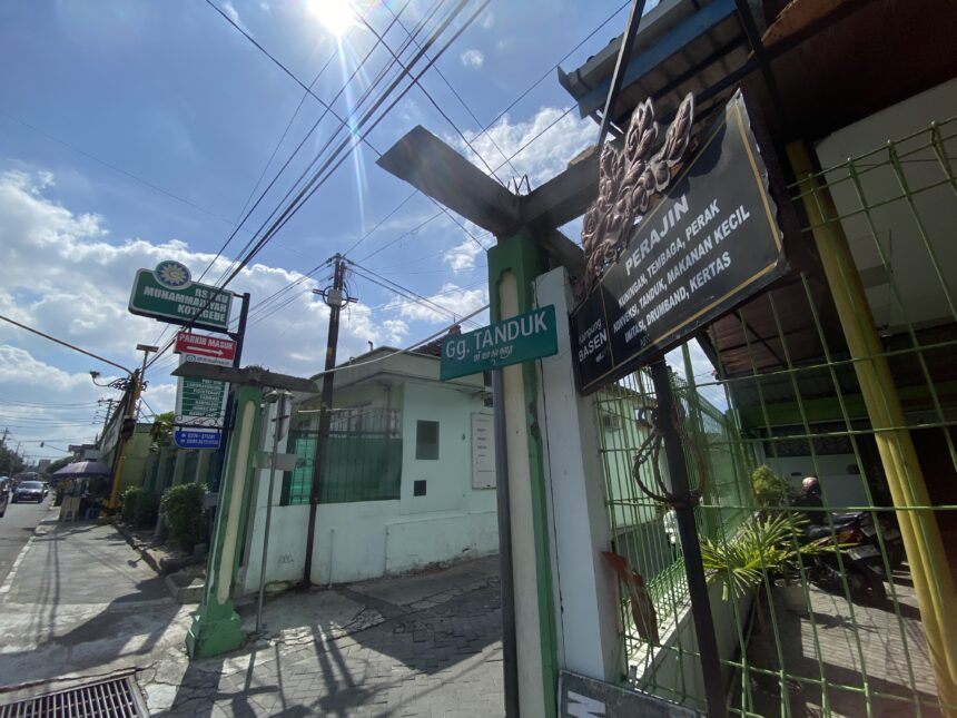 Sunlit urban street with overhead power lines, a green fence, and shop signs in Indonesian.
