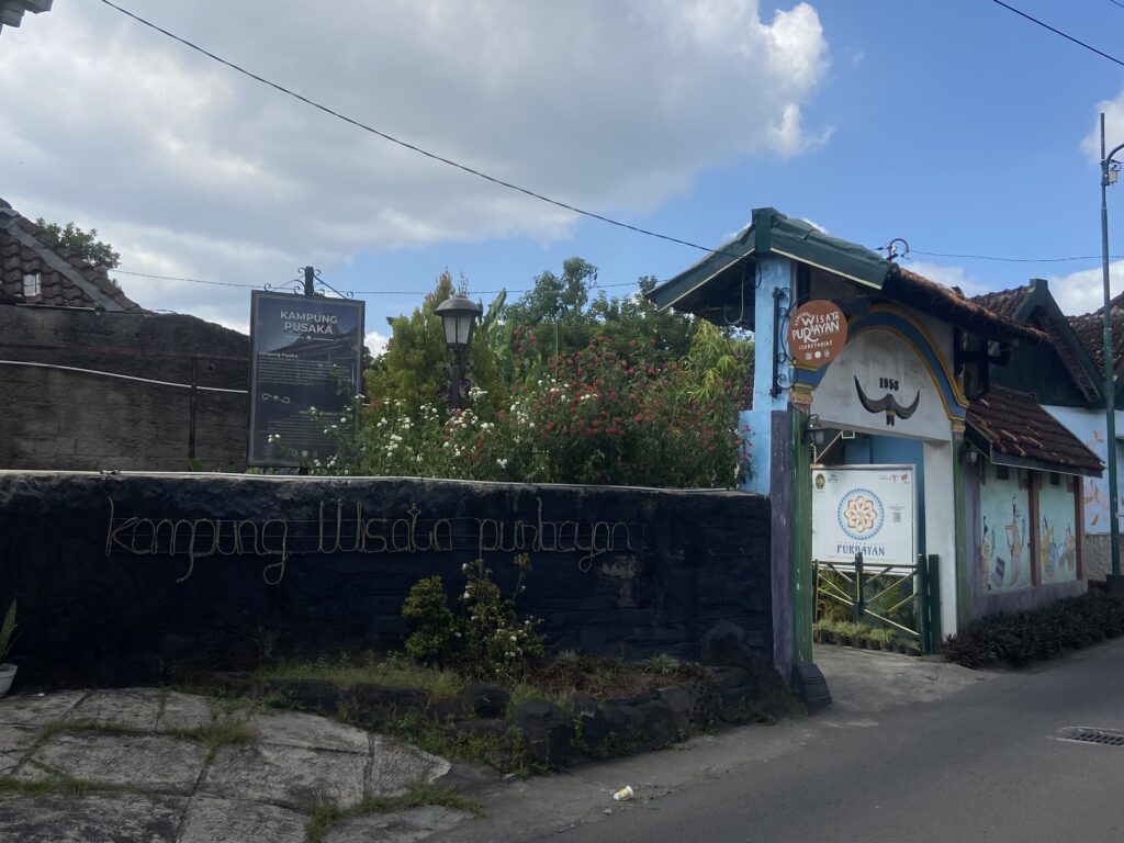 IMG_7351 - Mabur.co Entrance to a tourist area with a blue-and-green painted structure, orange circular sign reading'Wisata Purbayan Sekretariat', and an information board beside a black wall.
