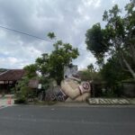 Street scene with a large decorative stone statue of a mythical creature at a roadside area, under a cloudy sky with green trees and a sign in Indonesian visible in the background.