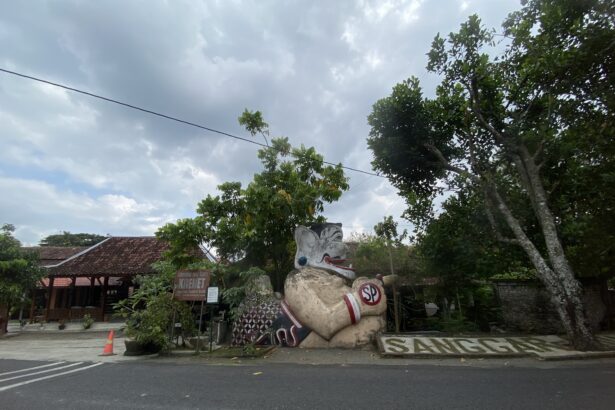 Street scene with a large decorative stone statue of a mythical creature at a roadside area, under a cloudy sky with green trees and a sign in Indonesian visible in the background.