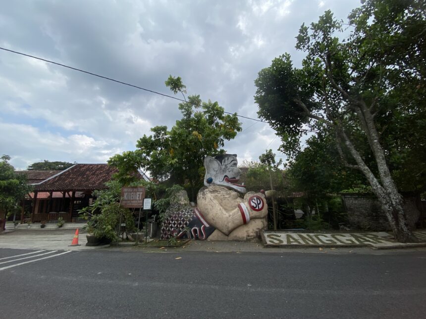 IMG_7402 - Mabur.co Street scene with a large decorative stone statue of a mythical creature at a roadside area, under a cloudy sky with green trees and a sign in Indonesian visible in the background.
