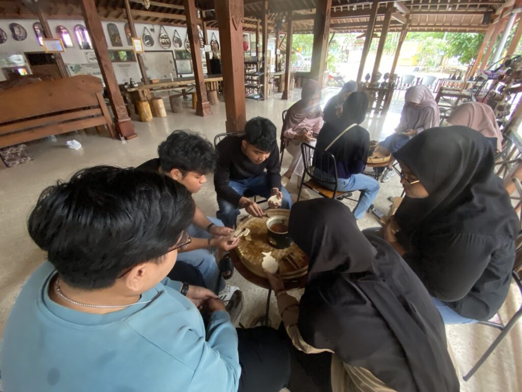 IMG_7410 - Mabur.co Group of people seated in a circle around a central pot, participating in a hands-on workshop inside a wooden, open-air hall.