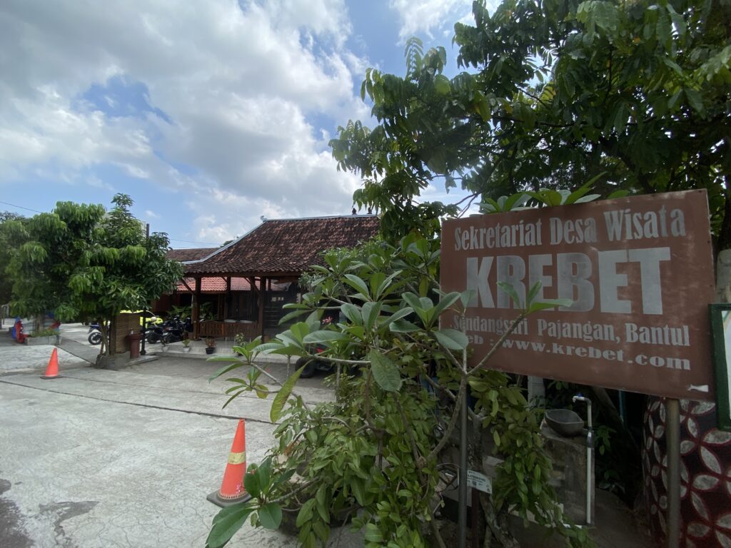 IMG_7433 - Mabur.co Outdoor scene with a large brown sign reading'Sekretariat Desa Wisata Krebet' beside greenery and a red-tiled building roof behind trees.