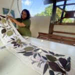 Woman in a green shirt unfolds a plant-printed quilt on a white table indoors, with an outdoor scene and wooden structure nearby