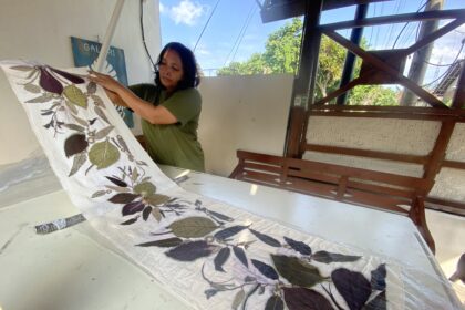 Woman in a green shirt unfolds a plant-printed quilt on a white table indoors, with an outdoor scene and wooden structure nearby