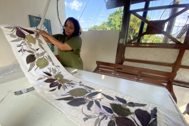 Woman in a green shirt unfolds a plant-printed quilt on a white table indoors, with an outdoor scene and wooden structure nearby