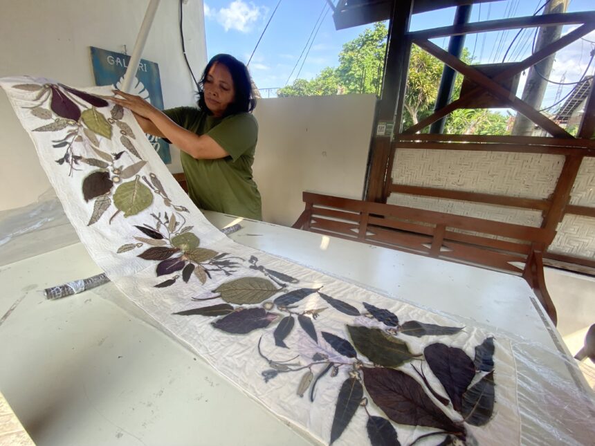 Woman in a green shirt unfolds a plant-printed quilt on a white table indoors, with an outdoor scene and wooden structure nearby