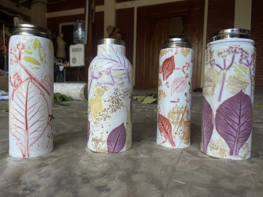 Row of four tall ceramic vases with botanical leaf patterns in pastel colors, placed on a concrete floor in a workshop setting.