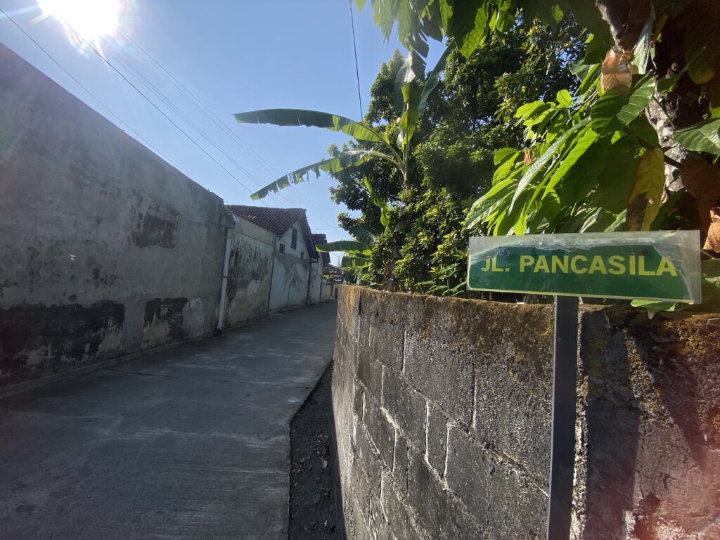Sunny alley with concrete walls and large banana leaves overhead; a green road sign reads 'JL PANCASILA' at right foreground