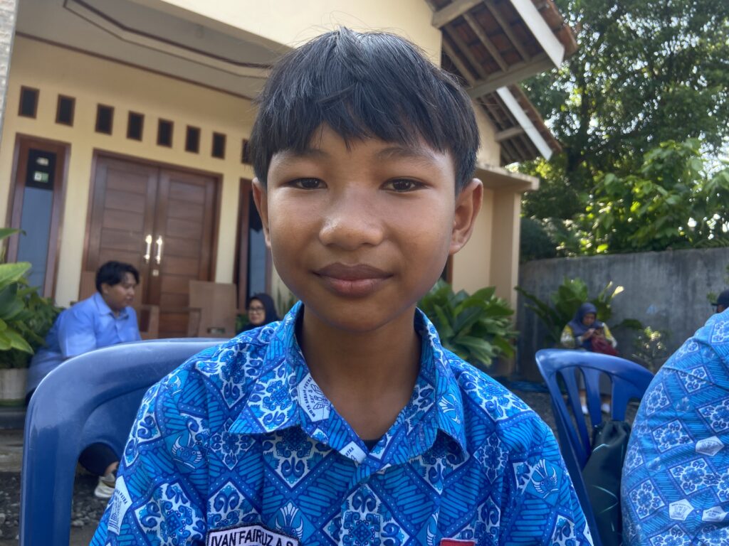 IMG_7486 - Mabur.co Close-up of a smiling boy in a blue patterned shirt, outdoors with a house and greenery in the background.