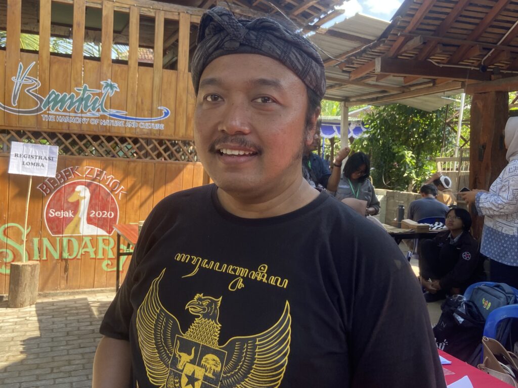 Close-up of a smiling man wearing a dark head wrap and a black T-shirt with a gold dragon design, standing in a sunny outdoor area with wooden walls and banners behind him.