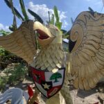 Colorful carved dragon or phoenix sculpture outdoors with yellow scales and outstretched wings, wearing a shield in a sunny garden.