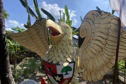 Colorful carved dragon or phoenix sculpture outdoors with yellow scales and outstretched wings, wearing a shield in a sunny garden.