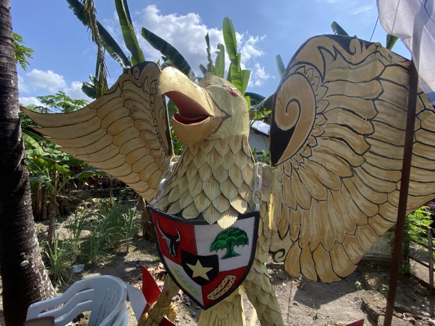 IMG_7497 - Mabur.co Colorful carved dragon or phoenix sculpture outdoors with yellow scales and outstretched wings, wearing a shield in a sunny garden.