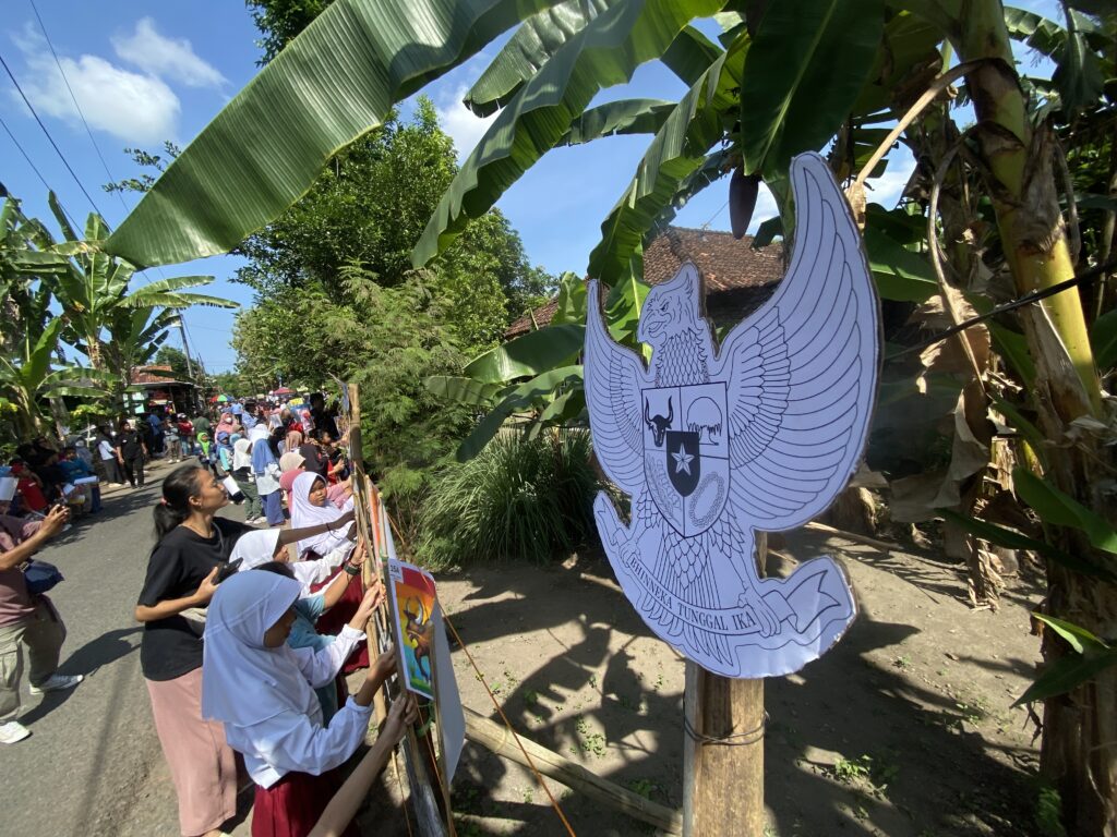 People line a street at a festival, holding posters, with a large Garuda emblem cutout hanging nearby among banana trees.