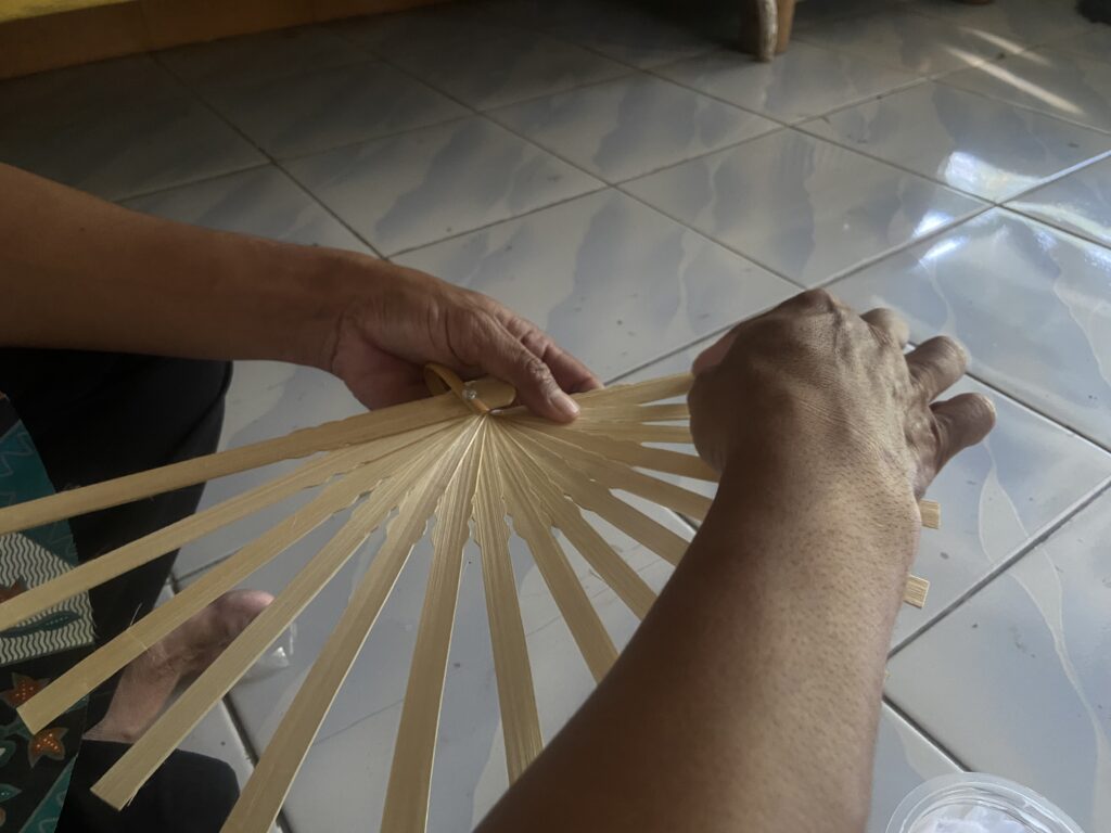 Hands weave a palm leaf fan, arranging thin slats in a radiating circle on a tiled floor.