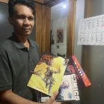 Man in a dark polo shirt holds up colorful comic books beside a wall calendar.