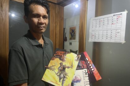 Man in a dark polo shirt holds up colorful comic books beside a wall calendar.