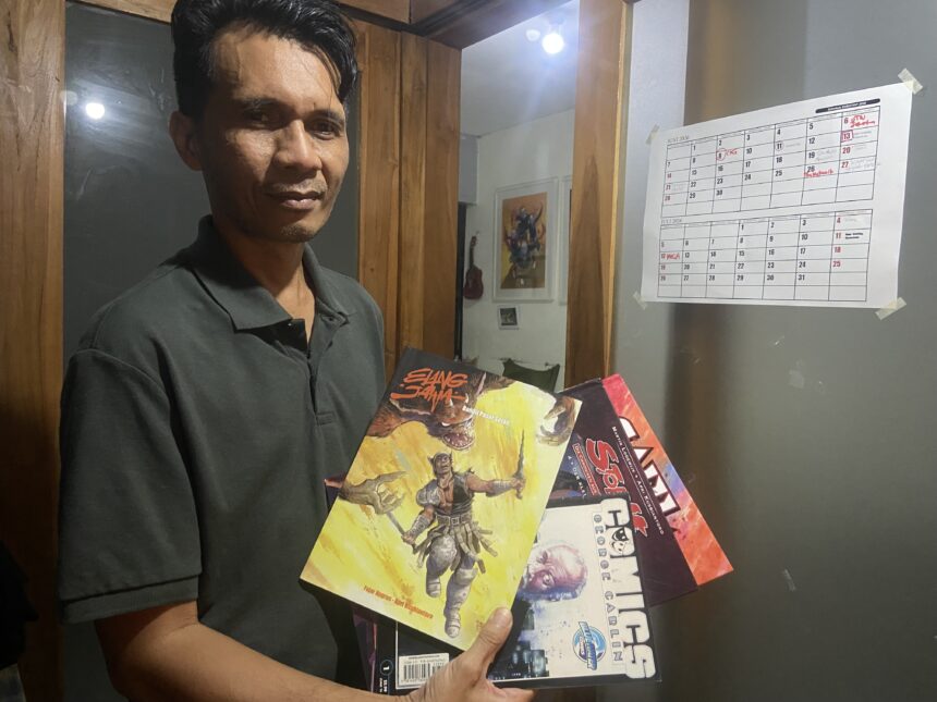 Man in a dark polo shirt holds up colorful comic books beside a wall calendar.