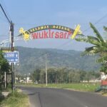 Curved welcome sign over a road reading 'Selamat Datang Wukirsari' with banana trees nearby and hills in the distance.