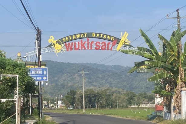 Curved welcome sign over a road reading 'Selamat Datang Wukirsari' with banana trees nearby and hills in the distance.