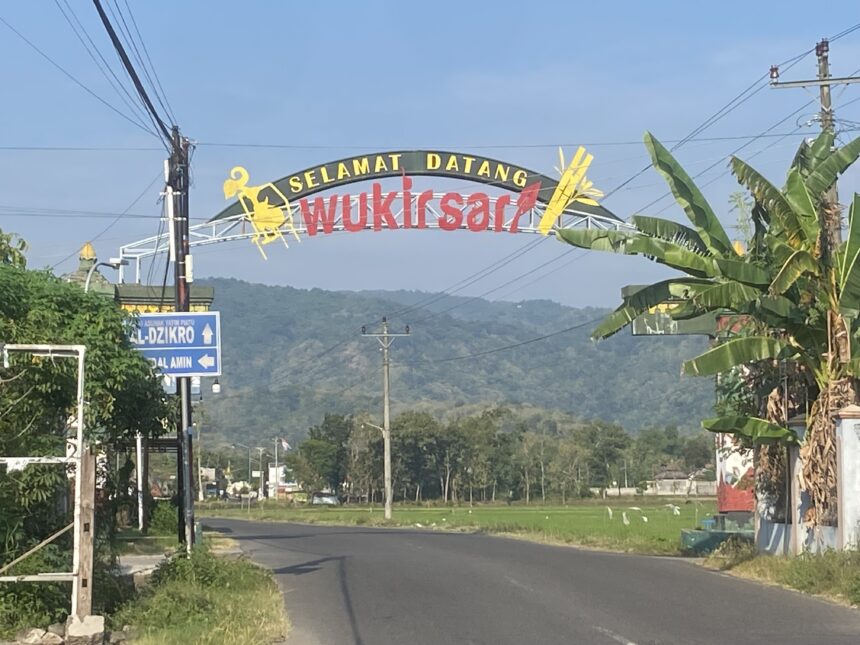 Curved welcome sign over a road reading 'Selamat Datang Wukirsari' with banana trees nearby and hills in the distance.
