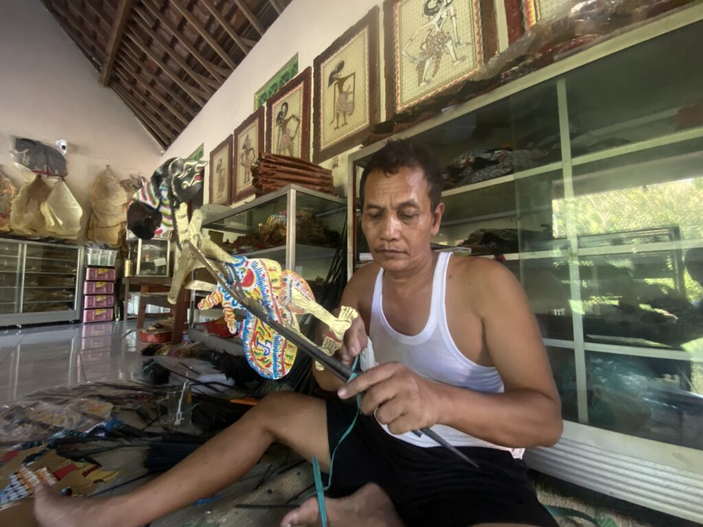 Man in a white tank top handcrafting a colorful decorative kite inside a shop with glass display cases nearby.