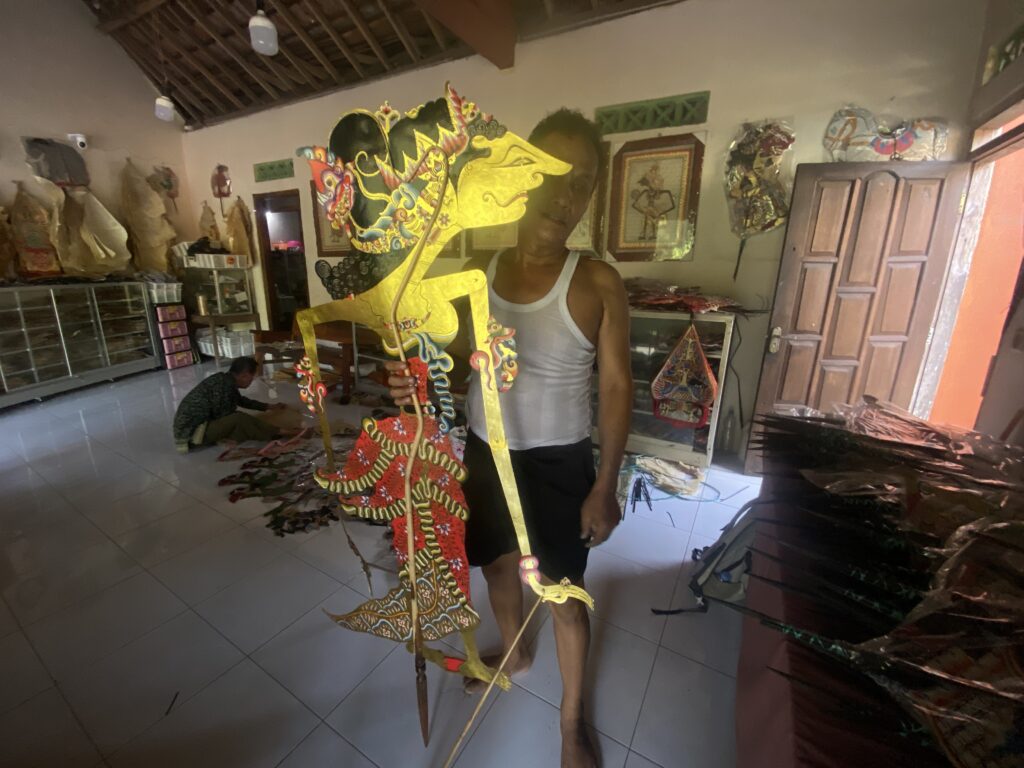 Man in a white tank top holding a large colorful dragon puppet inside a craft workshop.