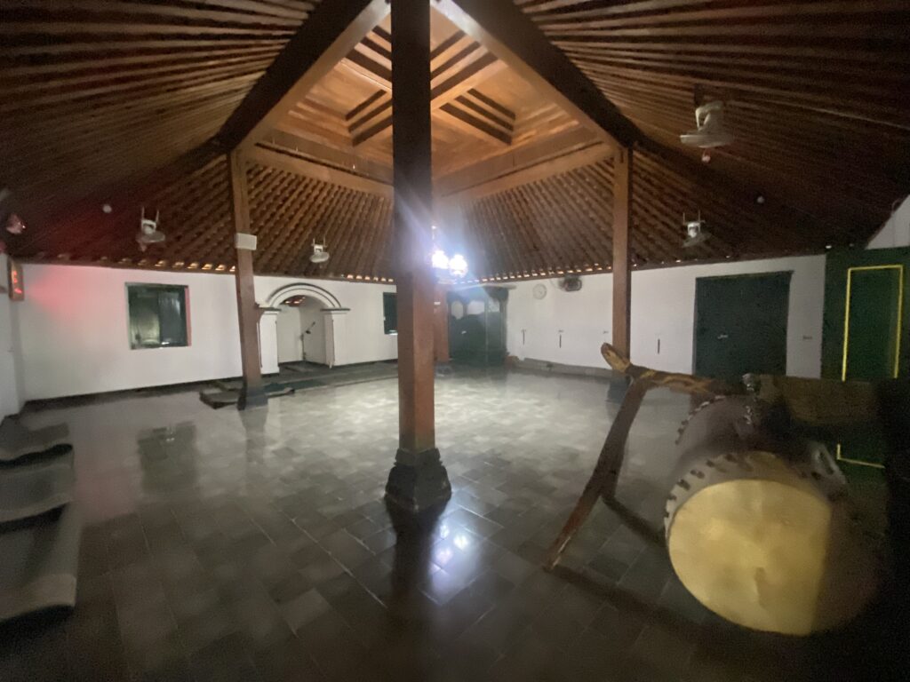 IMG_7777 - Mabur.co Interior view of a large wooden hall with a peaked ceiling and exposed beams, illuminated by a chandelier in the center. A circular drum sits in the foreground on the right, with tiled flooring and several pillars supporting the roof.