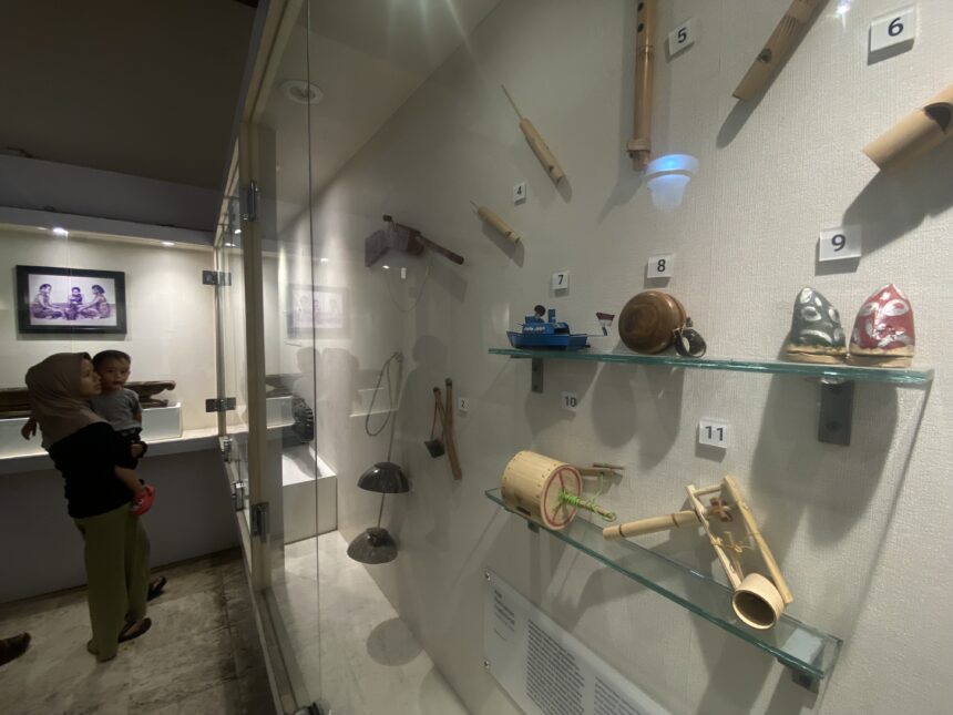 Museum display case with numbered wooden artifacts mounted on the wall; glass barrier reflects visitors in the foreground.