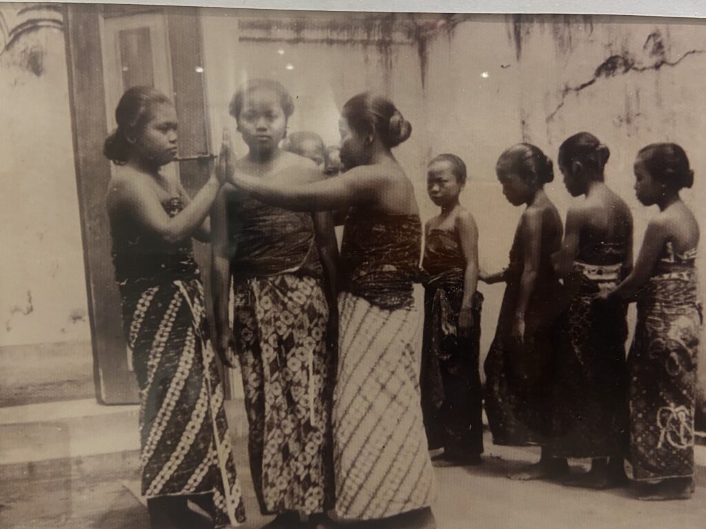 Vintage black-and-white photo of young women in patterned saris standing in a line outdoors; one resting a hand on another's shoulder.