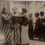 Vintage black-and-white photo of young women in patterned saris standing in a line outdoors; one resting a hand on another's shoulder.