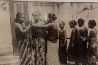 Vintage black-and-white photo of young women in patterned saris standing in a line outdoors; one resting a hand on another's shoulder.