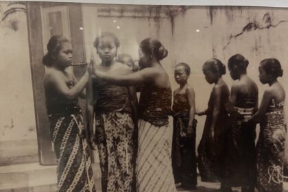 Vintage black-and-white photo of young women in patterned saris standing in a line outdoors; one resting a hand on another's shoulder.