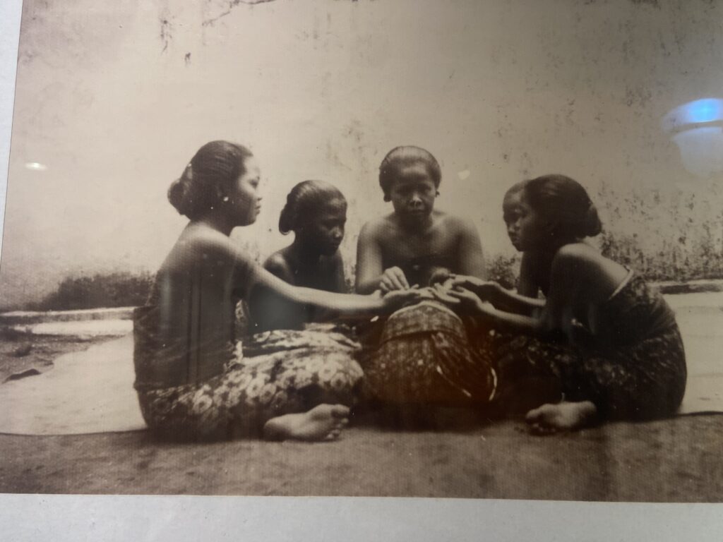 Black-and-white photo of four women sitting in a circle, heads close together, around a large woven basket on the ground.