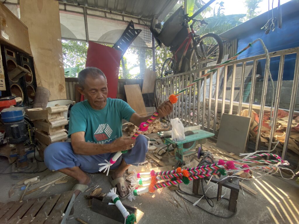 Older man in a teal shirt tying bright pom-poms onto a striped rope inside a cluttered workshop with wood and tools nearby.