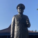 Bronze statue of a military officer in uniform and peaked cap, standing with a raised right hand against a bright blue sky.