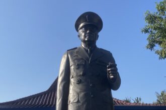 Bronze statue of a military officer in uniform and peaked cap, standing with a raised right hand against a bright blue sky.