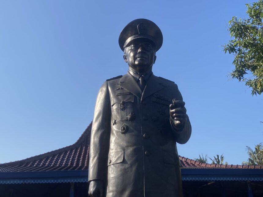 IMG_8041 - Mabur.co Bronze statue of a military officer in uniform and peaked cap, standing with a raised right hand against a bright blue sky.