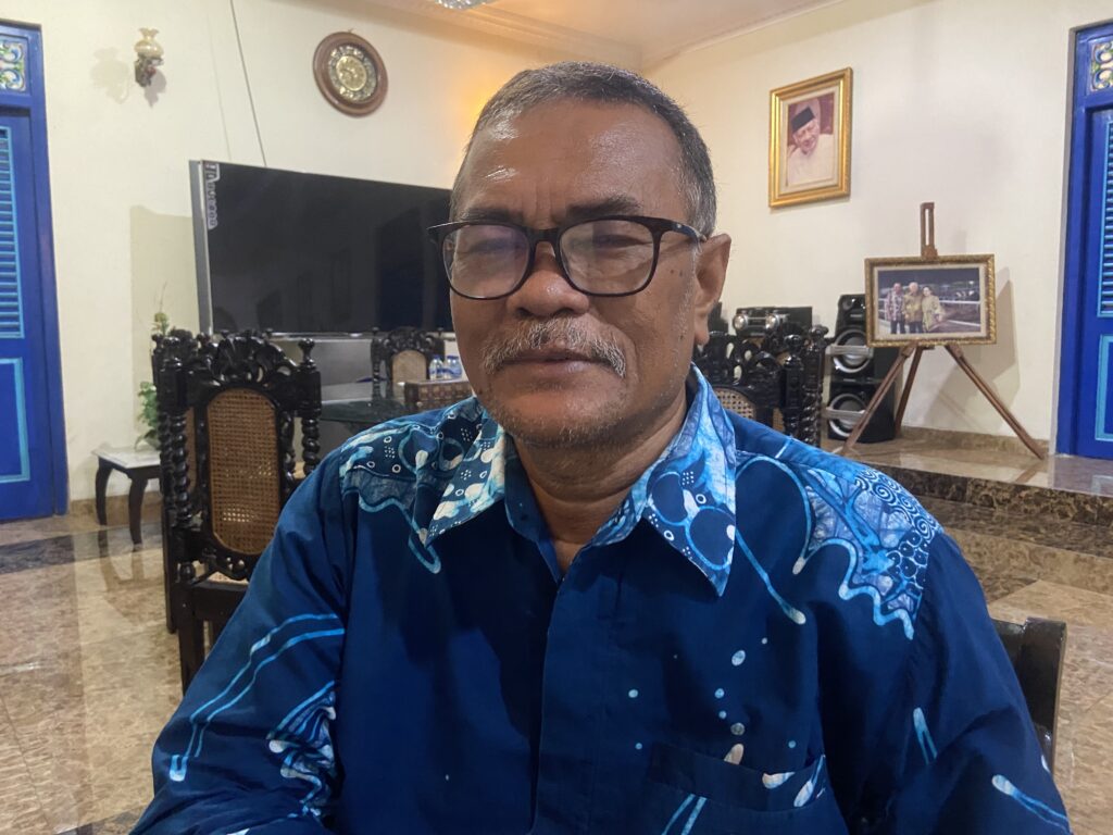 IMG_8050 - Mabur.co Selfie of a man with short gray hair and glasses wearing a blue patterned shirt, indoors with blue doors and framed photos behind him, including a portrait on the wall and a photo on an easel.