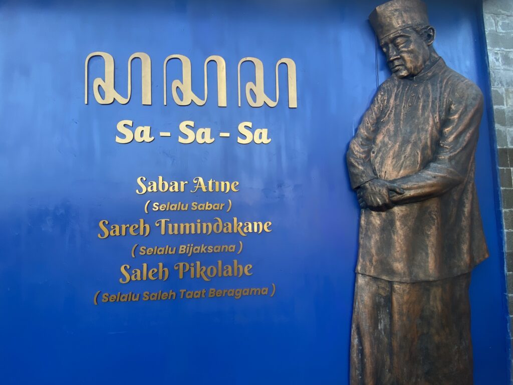 IMG_8057 - Mabur.co Bronze statue of a man in a traditional cap beside a blue wall with large gold Indonesian text on it.