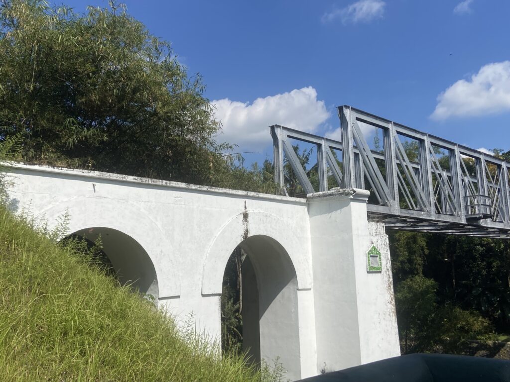 White wall with three arched openings sits beside a gray metal bridge frame; tall grasses and a bamboo tree loom to the left under a bright blue sky with clouds.
