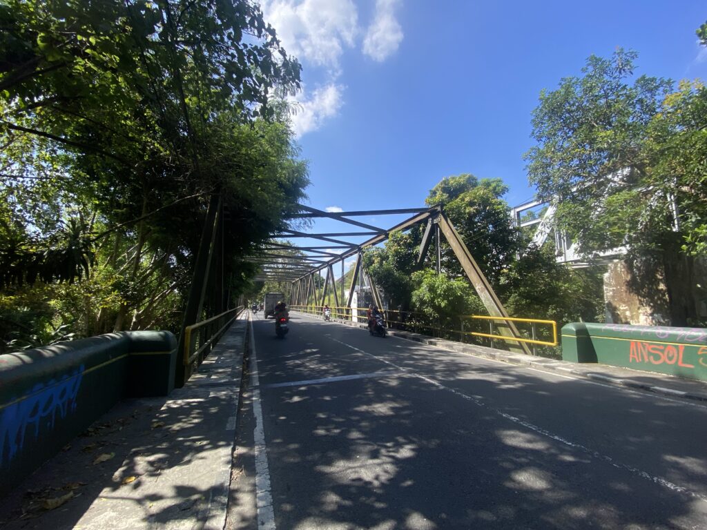 Bridge with metal truss supports over a tree-lined road; motorbikes ride beneath a bright blue sky.