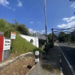 Street view with a metal pedestrian bridge, power pole, and lush green bushes under a blue sky with scattered clouds, on the right a roadway with scooters.