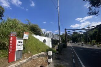 Street view with a metal pedestrian bridge, power pole, and lush green bushes under a blue sky with scattered clouds, on the right a roadway with scooters.